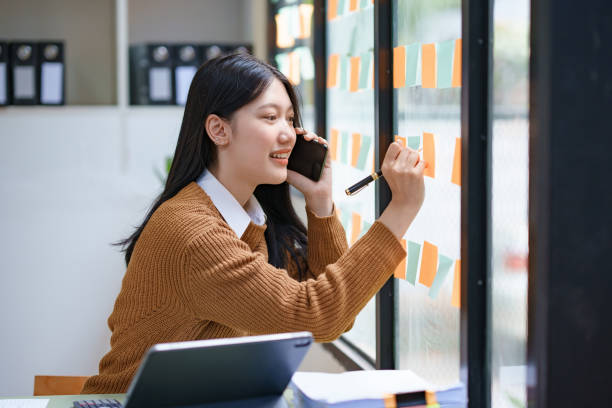 Young asian businesswoman multitasking, organizing sticky notes on a glass wall while talking on the phone in an office.