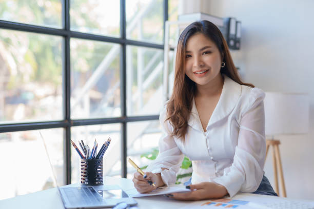 istock Professional asian businesswoman with a confident smile in a bright office setting with a laptop and documents. 2155123066