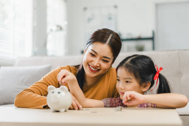 Cute little asian girl saving money by adding a coin in piggy bank with mother at home Cute little asian girl saving money by adding a coin in piggy bank with mother at home. savings account stock pictures, royalty-free photos & images