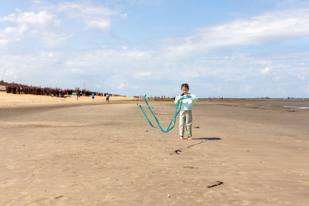 A little girl in glasses stands on an empty beach flying a kite.