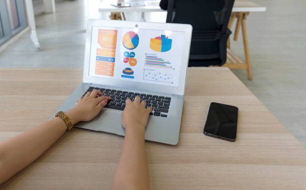 Back view of businesswoman sit at desk in office typing on laptop computer keyboard, making presentation with graph and multicolored chart.