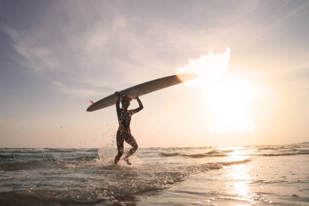 Female surfer wearing a swimsuit holding a surfboard on her head, entering out of sea after surfing on waves, silhouette in tropical golden light