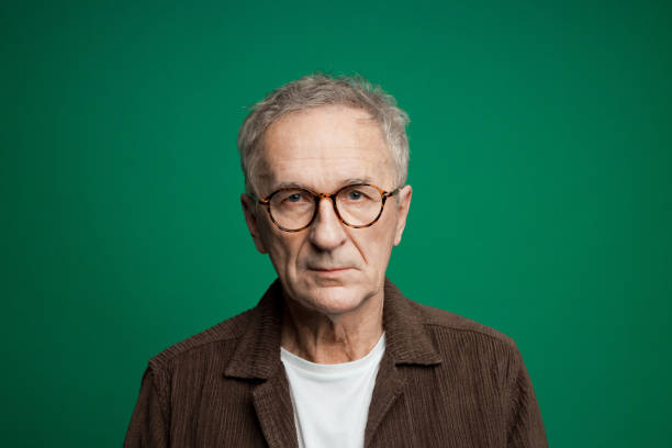 Sad senior man wearing white t-shirt and brown shirt looking at camera. Studio shot, green background.