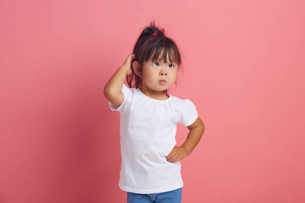 istock Confused little child scratches her head with thoughtful look has a puzzled expression dressed in neutral white t shirt looks with puzzlement aside, poses against pink background. 2155163747