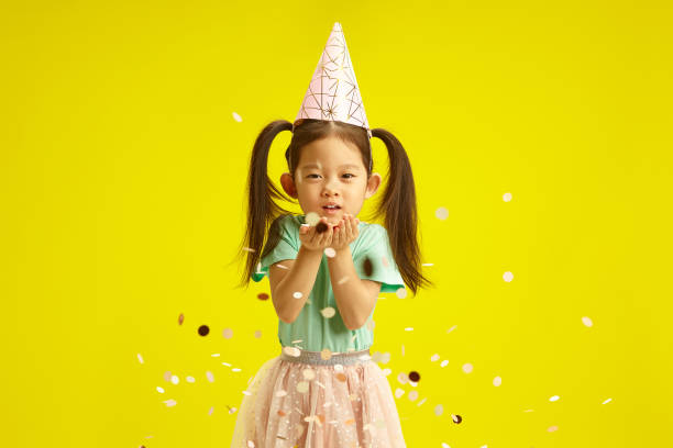istock Joyful Child with Conical Hat Blowing Confetti from hands, Celebrating Birthday Standing on Bright Yellow Background 2155164907