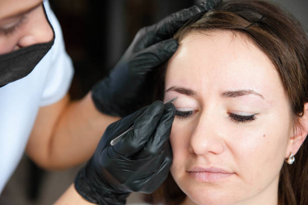 istock Browist makes eyebrow correction with tweezers. A model with dark hair in a light studio is sitting on an eyebrow correction at the eyebrow master. 2155180873