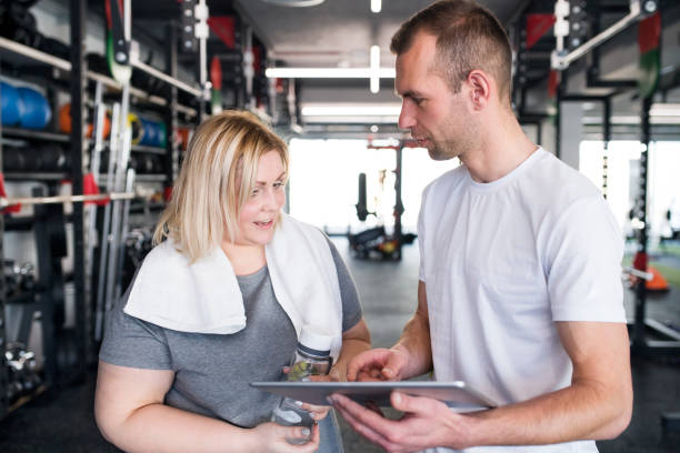 istock Overweight woman consulting with trainer, discussing daily workout plan. Personal trainer talking with client, discussing exercise program in gym. 2155182711