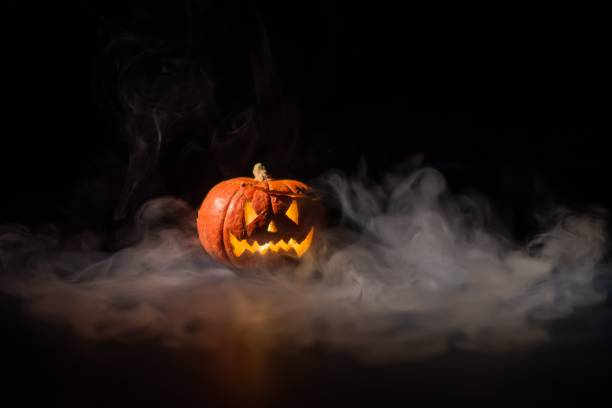 istock Halloween, orange pumpkin with a scary luminous face on a dark background. Thick gray smoke comes out and spreads across the black table. A close-up of a flashlight on the eve of all the saints 2155206559