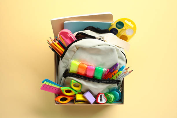 istock Open backpack filled with colorful school supplies such as pencils, markers, scissors, and notebooks in donation box on a pastel yellow background. Donation concept. 2155211064