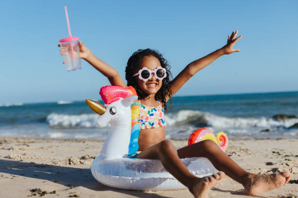 Happy girl on beach with unicorn float and drink enjoying summer vacation Cheerful young girl with sunglasses and unicorn float relaxing at the beach, holding a drink, and having fun during a summer vacation. black african girls pictures stock pictures, royalty-free photos & images