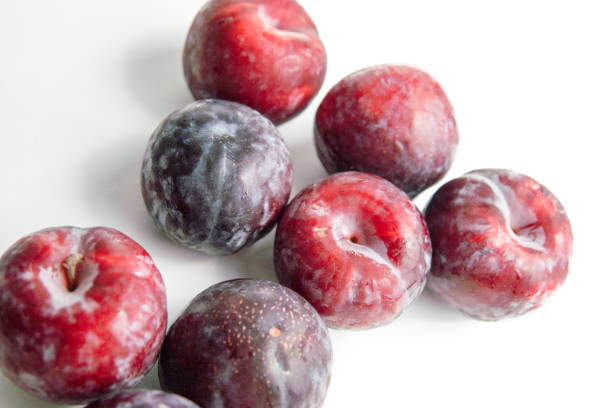 A close-up photograph of a cluster of fresh red plums with dewdrops, showcasing their vibrant colors and natural textures. The plums are arranged on a white surface with soft shadows in the background, emphasizing their juicy and appetizing appearance. This image is perfect for use in food photography, advertising, and editorial content, capturing the essence of fresh, organic fruit.