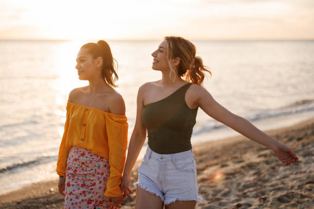 istock Female friends holding hands while walking together at the beach at sunset 2155244221