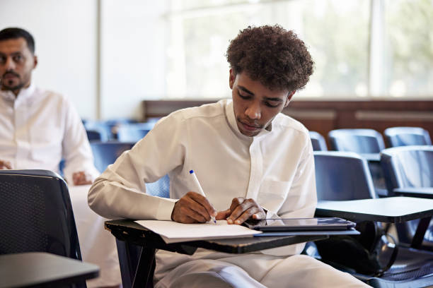 istock Focused male student writing in book while sitting on chair with digital tablet in classroom 2155267305