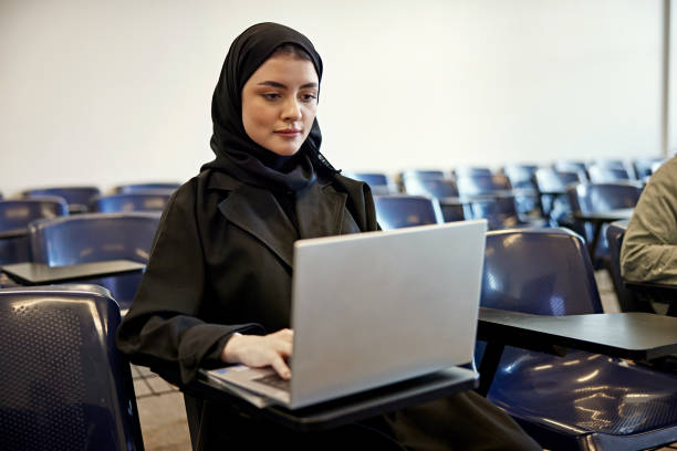 Focused young female student in hijab using laptop while sitting at desk in university classroom
