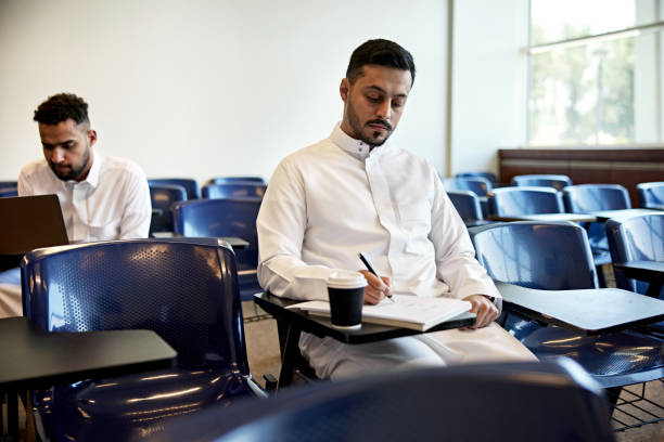 Young male student writing in book while sitting at desk in university classroom