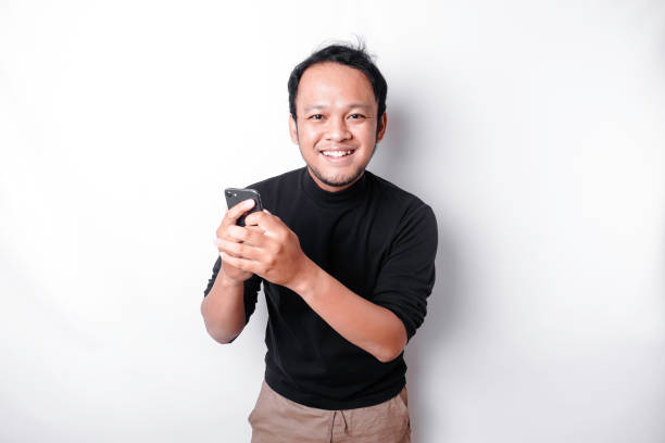 istock Excited Asian man wearing black shirt smiling while holding his phone, isolated by white background 2155273738
