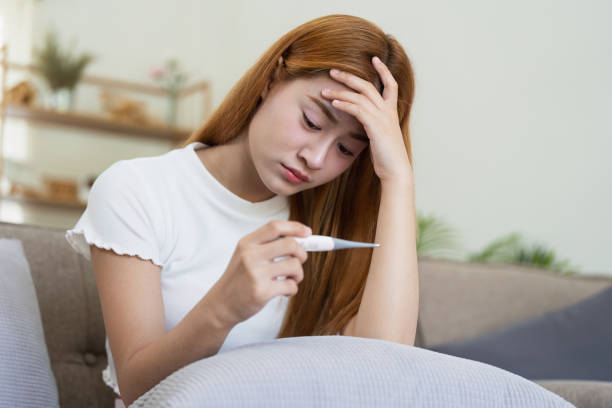 istock Sick young asian woman with thermometer suffering from cold on sofa, measuring body temperature with thermometer, touching forehead in sofa at home 2155274657