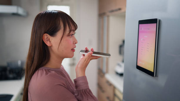 Asian woman checking ingredients on smart fridge screen and asking artificial intelligent smart phone app to get recipe ideas in kitchen