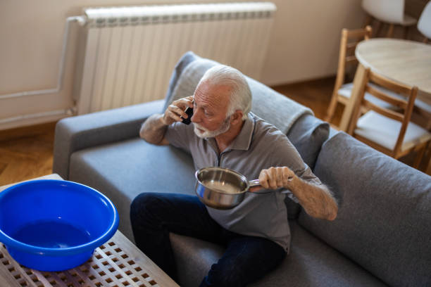 istock Worried senior man calling plumber while collecting water leaking from ceiling using utensil. Frustrated elderly man calling insurance about water leaks in the living room at home. 2155294664