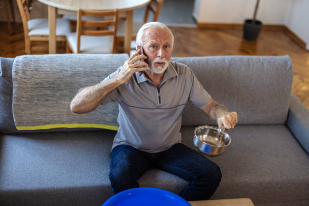 istock Worried senior man calling plumber while collecting water leaking from ceiling using utensil. Frustrated elderly man calling insurance about water leaks in the living room at home. 2155294768
