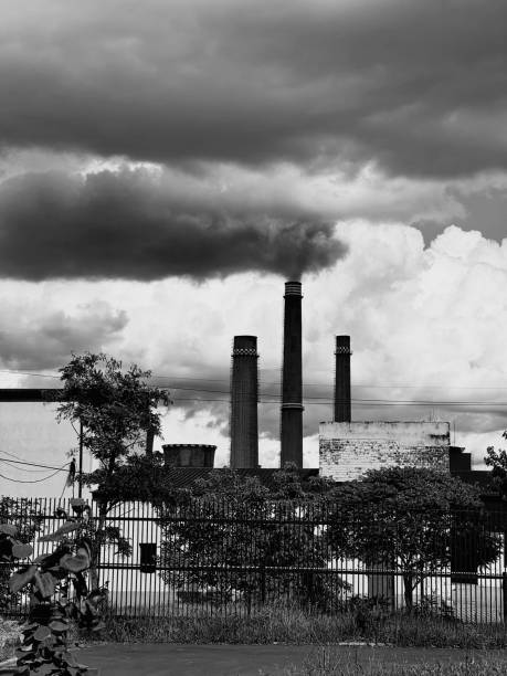 Black and white image of an industrial site with prominent smokestacks, set against a backdrop of dramatic clouds, evoking a sense of industrial history and environmental impact.