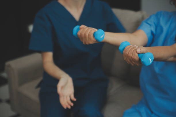 istock Old woman training with physiotherapist using dumbbells at home. Therapist assisting senior woman with exercises in nursing home. Elderly patient using dumbbells with outstretched arms. 2155311362