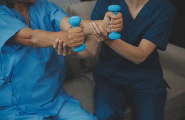 istock Old woman training with physiotherapist using dumbbells at home. Therapist assisting senior woman with exercises in nursing home. Elderly patient using dumbbells with outstretched arms. 2155311365