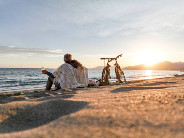 Mature couple enjoy drinks on beach Bicycles parked alongside. Mediterranean Sea and coastline distant, Liguria man 60s bicycle stock pictures, royalty-free photos & images