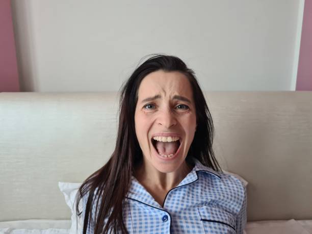 A woman is sitting up in bed, with a white headboard behind her, wearing a blue and white checkered pajama shirt, screaming