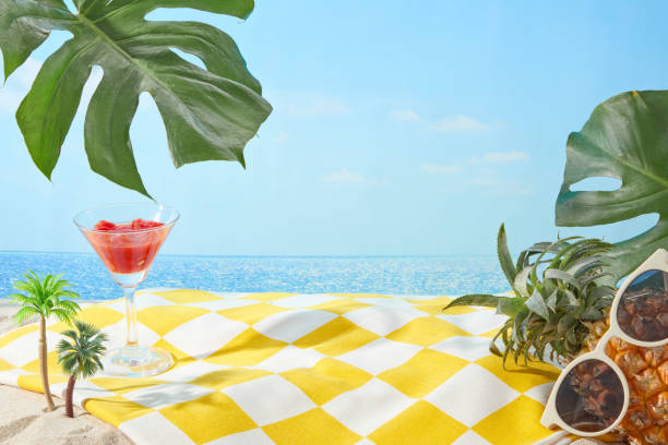 The yellow carpet with checkered pattern spread on sand beach side with a watermelon juice glass, pineapple and monstera leaves decorated above, over the sea and sky background. Vacant space in center