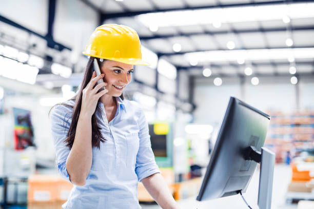 istock Female project manager standing in modern industrial factory by computer, making phone call. Manufacturing facility with robotics, robotic arms and automation. Storing products and materials in warehouse. 2155423754