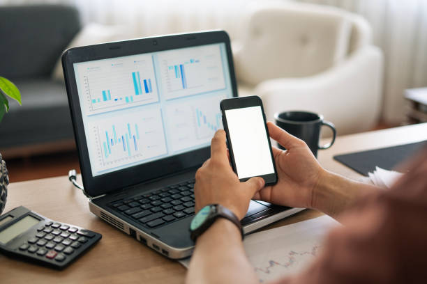 istock Close-up Young man using phone while reviewing financial reports on laptop at home , mock-up phone screen 2155427449
