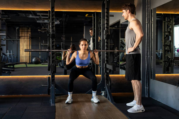 istock Young woman exercising with barbell while male trainer controlling lay during her workout at gym 2155453407
