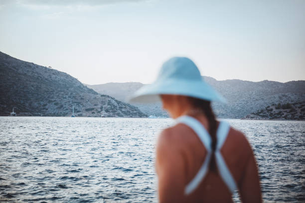 Soft focus rear view photo of a woman dressed in a nice sun hat on the beach enjoying beautiful sea view and mountains. Peaceful summer vacation.