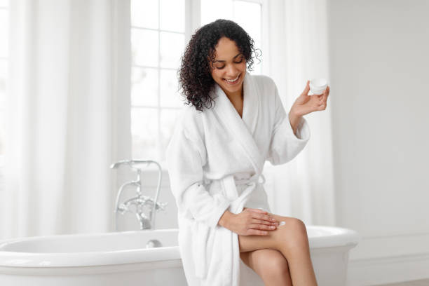 istock Young black lady applying moisturizer cream on legs, moisturizing skin after shower, doing aftershave skincare routine, sitting on bathtub in light bathroom at home 2155470667