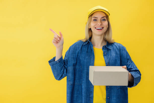 istock Smiling female courier in cap holding box and pointing on copy space with finger while looking at camera. Blond woman in jeans shirt bringing package and feeling excited over yellow background. 2155477192