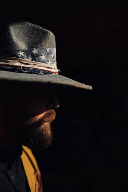 Profile view of a cowboy standing outside on sunny day. Close up of a young bearded man in stetson hat standing outdoors on sun. Cropped picture of a serious horse breeder standing outdoors in shadow.