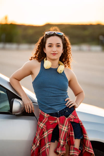 istock Curly-haired woman leaning on car with headphones, sunset backdrop. 2155482391