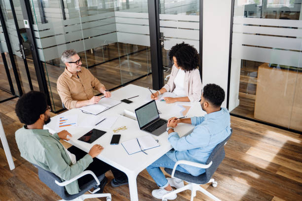 A team of four professionals are gathered around a white table in a modern office A team of four professionals are gathered around a white table in a modern office, indicative of a cooperative and strategic business meeting. Productive meetings and brainstorming concept constructive feedback stock pictures, royalty-free photos & images