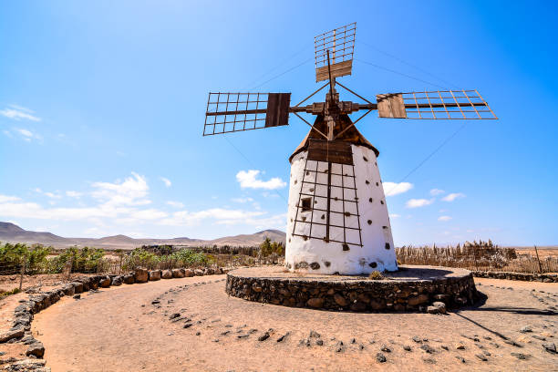 A windmill is standing in the middle of a desert. The sky is clear and blue, and the windmill is the only thing visible in the scene, image