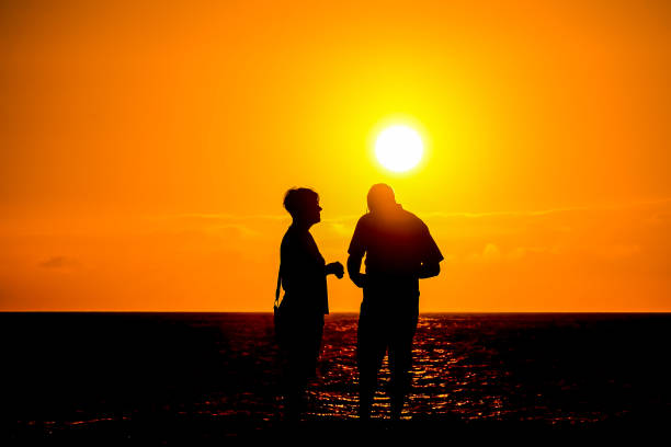 Two people are standing on the beach, looking out at the ocean. The sun is setting, casting a warm glow over the scene, image