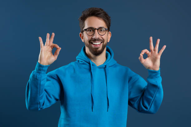 istock Young cheerful man in blue hoodie making OK sign with two hands 2155498479
