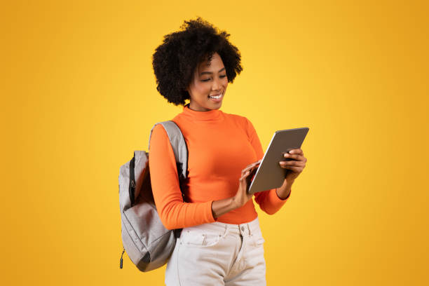 istock Engrossed African American woman with a delightful smile using a tablet, carrying a backpack 2155499574