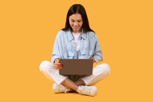 istock Young lady with laptop sitting cross-legged on yellow background 2155499995