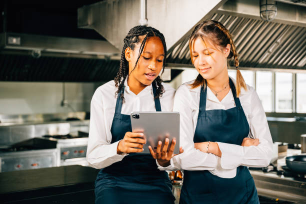 In a professional restaurant kitchen two famous chefs discuss video blog using a tablet. Modern technology aiding discussion colleagues smiling searching reading and connecting.