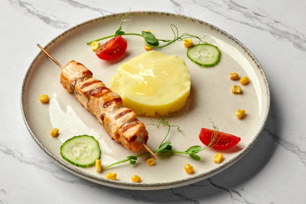 Delicious healthy food for kids menu. Mini grilled salmon skewer with portion of mashed potatoes garnished with fresh greens, corn kernels, cucumber and cherry tomato slices, against marble background