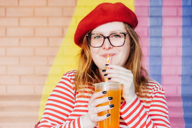 istock Smiling plus size woman in glasses and bright clothes drinking sugar flavored tapioca bubble tea near urban colorful painted brick wall. Happy fashionable hipster overweight girl. Street fashion 2155518323