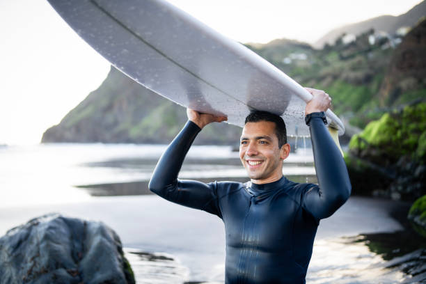 Back view of man with surfboard on beach. Rear view of surfer in wetsuit holding board and walking to sea waves at summertime. Surfing concept