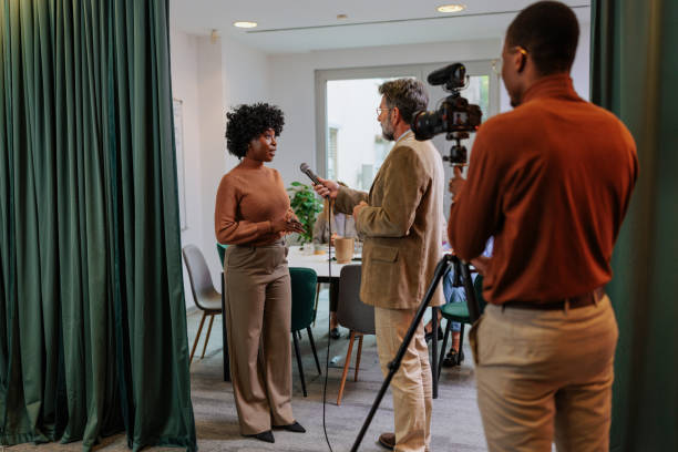 Young businesswoman sitting on sofa and talking to journalist during interview being filmed in modern office
