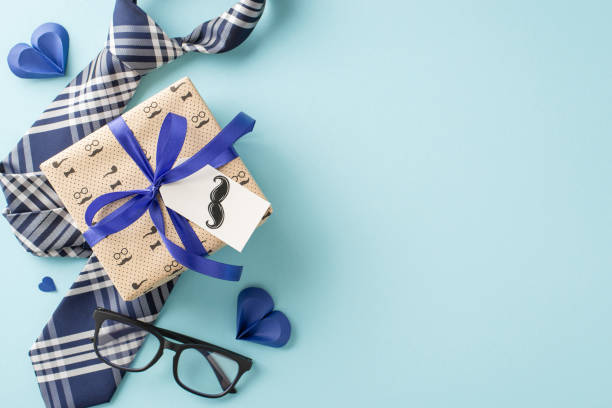Express your Father's Day sentiments uniquely with a top view composition featuring sleek necktie, gift box with creative mustache tag, glasses, and paper hearts on a gentle pastel blue base